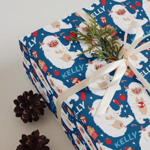 May include: Gift wrap featuring a repeating pattern of white yeti characters on a dark blue background. The yeti are holding various items. The word "KELLY" is printed in blue. A cream-colored ribbon and pine cones are also visible.