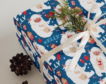 Puede incluir: Papel de regalo con un patrón repetido de personajes de yeti blancos sobre un fondo azul oscuro. Los yeti sostienen varios objetos. La palabra "KELLY" está impresa en azul. También se ven una cinta color crema y piñas.