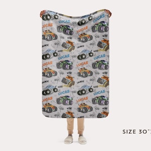 Op de afbeelding: Een witte babydeken met een herhalend patroon van monstertrucks in verschillende kleuren, waaronder oranje, blauw en groen. De trucks hebben de naam "Lucas" erop gedrukt. De deken is 76 cm bij 102 cm.