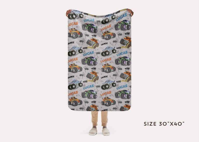 Op de afbeelding: Een witte babydeken met een herhalend patroon van monstertrucks in verschillende kleuren, waaronder oranje, blauw en groen. De trucks hebben de naam "Lucas" erop gedrukt. De deken is 76 cm bij 102 cm.
