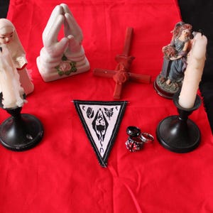 K&ouml;nnte beinhalten: Ein rotes Tuch zeigt verschiedene Gegenst&auml;nde, darunter eine Statue einer Frau, die eine Kerze h&auml;lt, ein Paar betende H&auml;nde, ein Kreuz, eine Statue einer Frau, ein Aufn&auml;her mit einem Symbol und mehrere Ringe. Die Gegenst&auml;nde sind auf einer roten Oberfl&auml;che angeordnet.