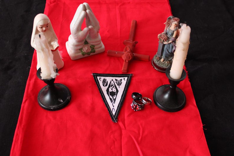 K&ouml;nnte beinhalten: Ein rotes Tuch zeigt verschiedene Gegenst&auml;nde, darunter eine Statue einer Frau, die eine Kerze h&auml;lt, ein Paar betende H&auml;nde, ein Kreuz, eine Statue einer Frau, ein Aufn&auml;her mit einem Symbol und mehrere Ringe. Die Gegenst&auml;nde sind auf einer roten Oberfl&auml;che angeordnet.