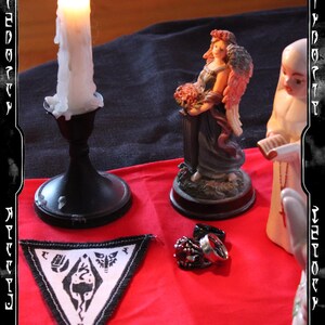 K&ouml;nnte beinhalten: Eine brennende wei&szlig;e Kerze in einem schwarzen Kerzenhalter, eine kleine Statue einer Frau mit Engelsfl&uuml;geln, ein Aufn&auml;her mit einem schwarz-wei&szlig;en Design und Ringe auf einer roten Oberfl&auml;che. Die Szene ist mit einem schwarzen Rahmen versehen.