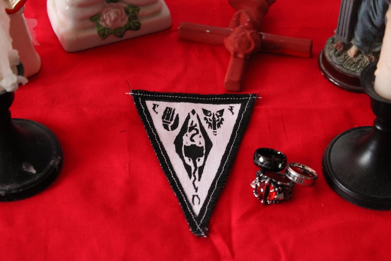 K&ouml;nnte beinhalten: Ein wei&szlig;-schwarzer, dreieckiger Aufn&auml;her mit Symbolen, umrandet von einem schwarzen Rand, liegt auf einer roten Oberfl&auml;che. Mehrere Ringe, darunter ein Ring mit rotem Edelstein, befinden sich in der N&auml;he. Ein Holzkreuz und andere dekorative Gegenst&auml;nde sind im Hintergrund.