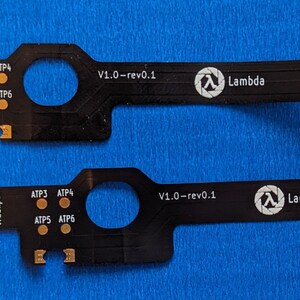 Op de afbeelding: Twee zwarte printplaten met gouden connectoren en cirkelvormige uitsparingen. Elke printplaat heeft de tekst "P2 Trackpad", "ATP3 ATP4", "ATP5 ATP6", "V1.0-rev0.1" en het Lambda-logo. De platen liggen op een blauw oppervlak.