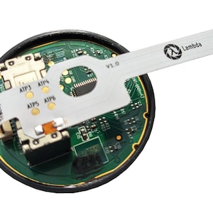 Puede incluir: Una placa de circuito circular con detalles verdes y dorados, con un cable de cinta blanco que se extiende hacia afuera. El cable tiene el logotipo de Lambda y el texto "Lambda" impreso. La placa tiene varios componentes y el texto "Trackpad" es visible.