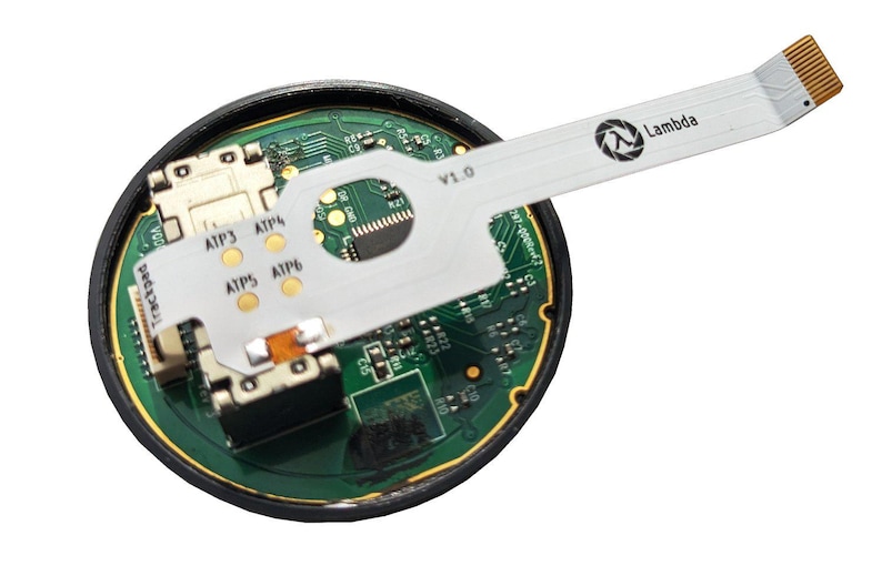 Op de afbeelding: Een ronde printplaat met groene en gouden accenten, met een witte lintkabel die naar buiten uitsteekt. De kabel heeft het Lambda-logo en de tekst "Lambda" erop gedrukt. De printplaat heeft verschillende componenten en de tekst "Trackpad" is zichtbaar.