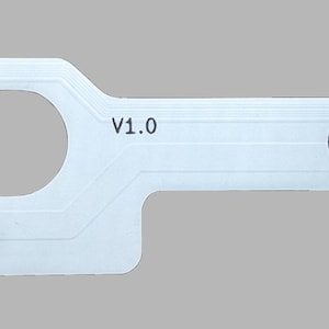 Op de afbeelding: Een witte printplaat met een trackpad-ontwerp, met de tekst "P2 Trackpad" en "Lambda". Het heeft een cirkelvormige opening en verschillende goudkleurige contactpunten. De printplaat heeft een rechthoekige vorm met twee verlengde uiteinden.