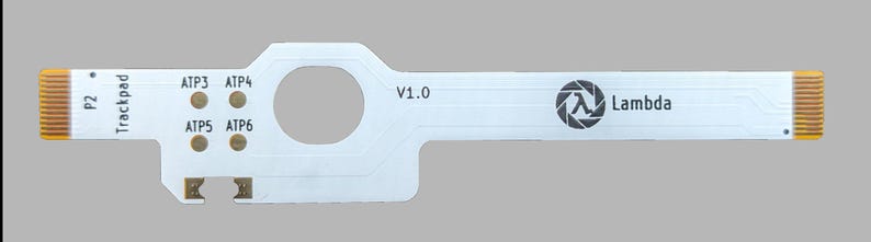 Op de afbeelding: Een witte printplaat met een trackpad-ontwerp, met de tekst "P2 Trackpad" en "Lambda". Het heeft een cirkelvormige opening en verschillende goudkleurige contactpunten. De printplaat heeft een rechthoekige vorm met twee verlengde uiteinden.