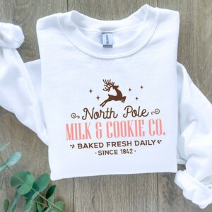 May include: White sweatshirt with a brown reindeer graphic and the text "North Pole MILK & COOKIE CO. BAKED FRESH DAILY SINCE 1842." The sweatshirt is made of a soft material.
