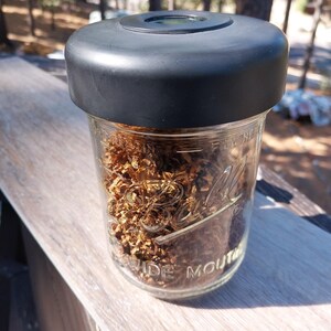 May include: A clear glass jar with a black lid, filled with dried, shredded brown material. The jar is labeled "WIDE MOUTH" in embossed lettering. The jar sits on a wooden surface, with a blurred forest background.
