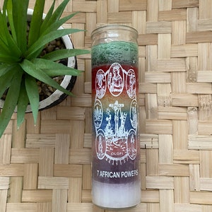 May include: A glass candle with a multi-colored design featuring seven saints and the text "7 AFRICAN POWERS". The candle is on a woven surface.