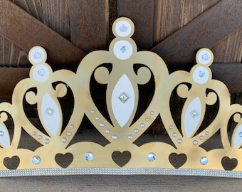 May include: A gold crown with white accents and rhinestones. The crown has a heart-shaped base and a row of rhinestones along the bottom.