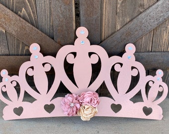 May include: A pink glitter princess crown with a decorative floral design. The crown is made of wood and has a cutout design.