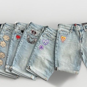 May include: A collection of light-wash denim shorts with various embroidered patches. Patches include roses, hearts, stars, and other designs. The shorts are arranged in a row, showcasing different styles and embellishments.