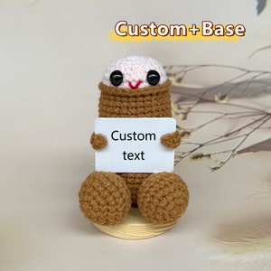 Puede incluir: Una figura amigurumi de crochet con un cuerpo marr&oacute;n y una cabeza rosa. La figura sostiene un cartel blanco que dice "Custom text".