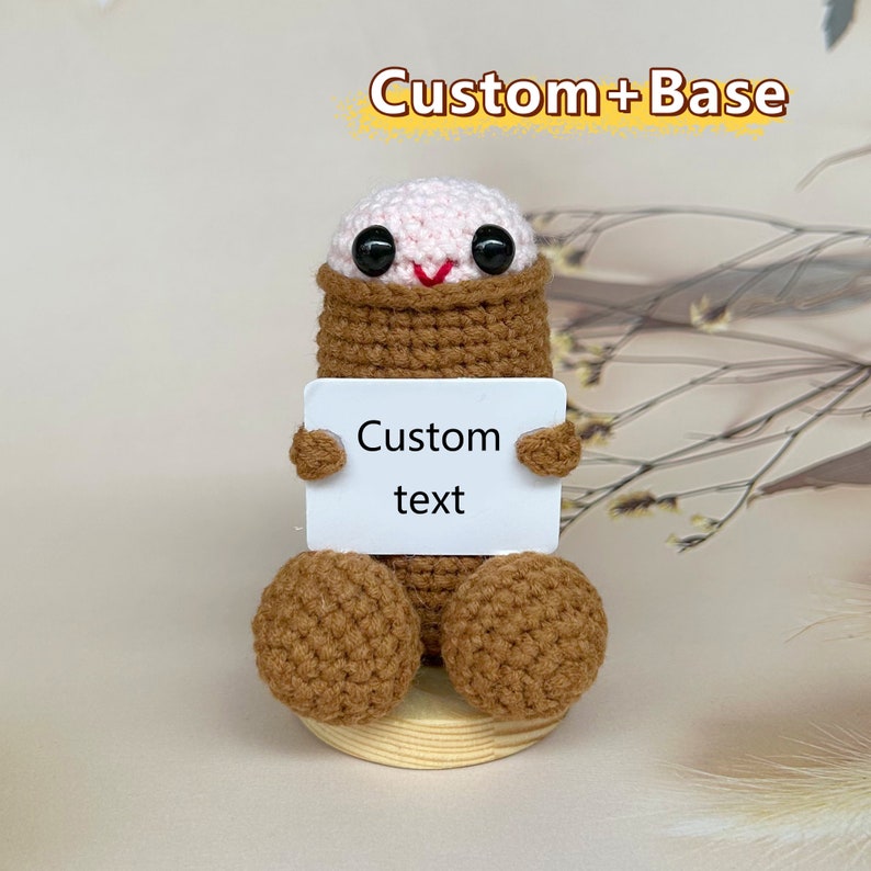 Puede incluir: Una figura amigurumi de crochet con un cuerpo marr&oacute;n y una cabeza rosa. La figura sostiene un cartel blanco que dice "Custom text".