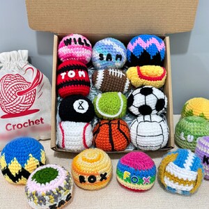 May include: A collection of small, round, crocheted yarn balls in a cardboard box and on a surface. The balls feature various patterns, including sports themes like a football, basketball, and soccer ball, and some are personalized with names like WILL, SAM, TOM, ROX, and FOX.