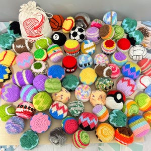 May include: A collection of small, round, crocheted yarn balls in various colors and patterns, including sports-themed designs like basketballs, soccer balls, and footballs, alongside a white drawstring storage bag with a red yarn logo.