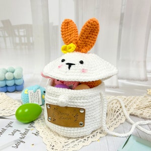 May include: White crocheted bunny basket with orange ears and a yellow flower. The basket is open and filled with colorful Easter eggs. A brown leather tag with the text "Penny's Easter" is attached to the front of the basket.