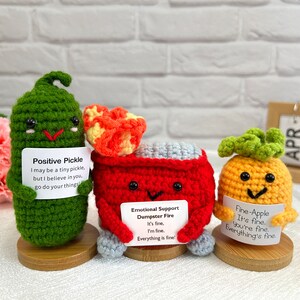 May include: Three crocheted plushies with encouraging messages. A green pickle plush says "Positive Pickle I may be a tiny pickle, but I believe in you, go do your things!" A red fire truck plush says "Emotional Support Dumpster Fire It's fine, I'm fine. Everything is fine!" A yellow pineapple plush says "Fine-Apple It's fine. You're fine. Everything's fine."