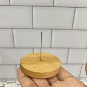 Puede incluir: Una base de madera con un pin met&aacute;lico en el centro. La base es de color marr&oacute;n claro y tiene una superficie lisa.