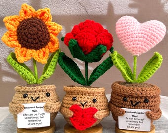 May include: Three crocheted potted plants with motivational messages. The plants are a sunflower, a red rose, and a pink heart. Each pot has a happy face and is holding a small heart. The message on the cards reads "Emotional Support Plant. Life can be tough sometimes, but remember so are you!"