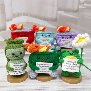 May include: A collection of handmade crochet figures, including a red crab, a green pea pod, a green frog, and others. Each figure holds a small sign with humorous phrases like "Emotional Support Dumpster Fire." The figures are on wooden bases.