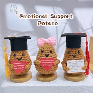 May include: Three crocheted potato figures wearing graduation caps and holding signs with encouraging messages. The signs read "Emotional Support Potato I may be a tiny potato, but I believe in you. Go do your thing!" and "Pocket Hug Potato I'm here to remind you that you're doing great and you are not alone. I'm here for you. You've got this!"