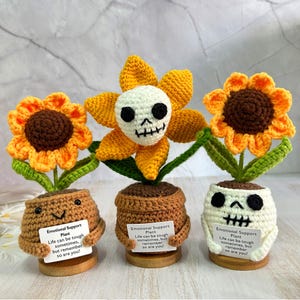 May include: Three crocheted sunflower plants with smiling faces and skull faces. Each plant has a sign that reads "Emotional Support Plant. Life can be tough sometimes, but remember so are you!"