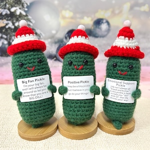 May include: Three crocheted pickle ornaments wearing Santa hats and holding signs with positive messages. The signs read "Big Fan Pickle", "Positive Pickle", and "Emotional Support Pickle".
