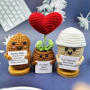 May include: Three crocheted bean-shaped figures holding signs with positive messages. The figure on the left is brown and says "Positive Bean I may be a tiny bean, but I believe in you, Go do your thing!" The middle figure is brown with a green plant and a red heart on top and says "My heart beats for you!!" The figure on the right is white with a brown top and says "I can't espresso how much you mean to me!"