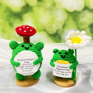 May include: Two crocheted green frog figurines with white faces and black eyes. The frog on the left is wearing a red and white mushroom hat and holding a sign that reads "Emotional Support Frog Life can be tough sometimes, but remember so are you!" The frog on the right is holding a sign that reads "Emotional Support Frog Be like frogs, just eat what bugs you."