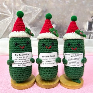 May include: Three handmade crochet pickle ornaments wearing Santa hats. Each green pickle has a smiling face and holds a sign with a different message. The ornaments sit on small wooden bases, ideal for festive decoration.