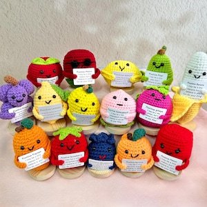 May include: A collection of 14 crocheted fruit plushies, each with a different fruit design and a positive message. The fruits include a grape, lemon, peach, papaya, strawberry, blueberry, apricot, melon, banana, pear, and apple. Each plush is holding a small sign with a message about believing in yourself and doing your thing.