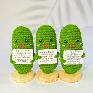 May include: Three green crocheted pickle figurines with smiling faces and different messages on cards. The messages are "Big Fan Pickle", "Positive Pickle", and "Emotional Support Pickle".