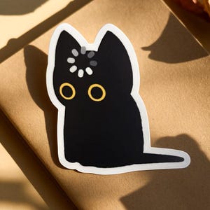 May include: A black cat sticker with a white border. The cat has large yellow eyes and a small design on its head. The sticker is on a brown surface.