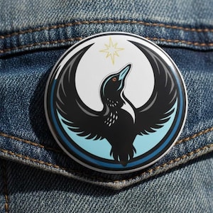 Pode incluir: Um botão redondo com um desenho estilizado de um pássaro. O pássaro é preto com as asas abertas, sobre um fundo branco com uma borda azul clara. Uma estrela amarela está acima da cabeça do pássaro. O botão está preso a uma jaqueta jeans.