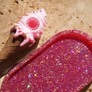 Pode incluir: Um cachimbo de silicone em forma de cone de sorvete rosa e marrom com uma bandeja de glitter rosa. O cachimbo tem uma tigela de vidro transparente e é decorado com granulado.