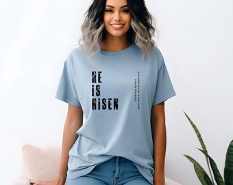 May include: A light blue t-shirt with the words "HE IS RISEN" in a distressed black font. The shirt also has text on the right side that reads "MATTHEW 28:6 HE IS NOT HERE". The shirt is made of a soft material.