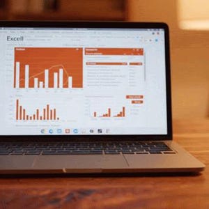 Op de afbeelding: Een laptop met een spreadsheet met grafieken in oranje en wit. Het scherm toont datavisualisaties. De laptop staat op een houten bureau, met een lamp en een boekenkast op de achtergrond.