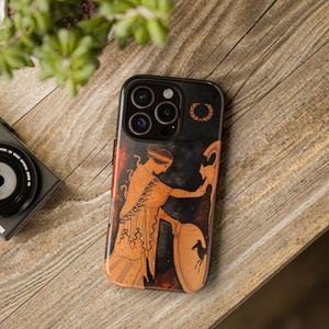 May include: Black phone case with an ancient Greek illustration in orange and black. The design shows a figure with a shield and spear. A laurel wreath is at the top. The case is on a wooden surface.