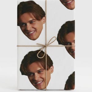 May include: White gift wrap featuring a repeating pattern of a smiling face with brown hair. The package is tied with twine and a bow.