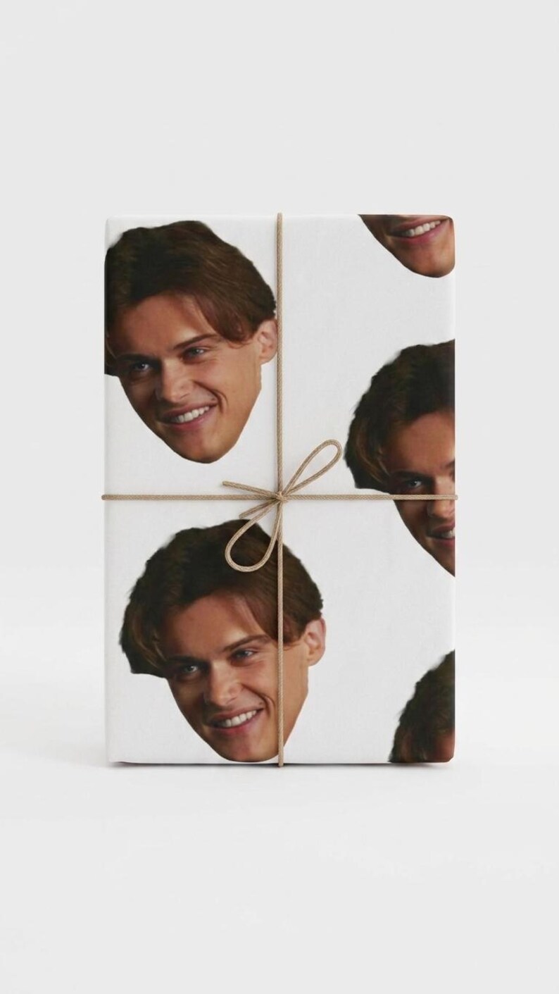 May include: White gift wrap featuring a repeating pattern of a smiling face with brown hair. The package is tied with twine and a bow.
