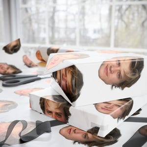 May include: White gift boxes wrapped in paper printed with a person's face. The boxes are stacked, with the faces visible on the top and sides. Scattered face cutouts and a pair of scissors are on the table.