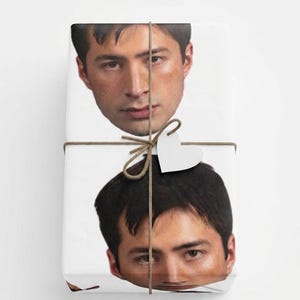 May include: Gift-wrapped package featuring a repeating pattern of a person's face. The wrapping paper is white, and the package is tied with twine and a heart-shaped tag. The overall aesthetic is simple and modern.