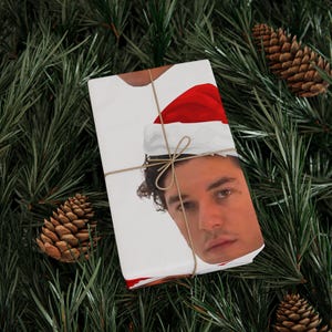 May include: A rectangular gift box wrapped in white paper featuring a printed graphic of a person wearing a red and white Santa hat. The package is tied with thin, light brown twine, creating a simple and clean presentation for the holiday season.