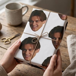 May include: A wrapped gift with a white paper featuring repeating images of a smiling person's face. The gift is tied with twine and held by hands. A pair of gold scissors and a mug are in the background.