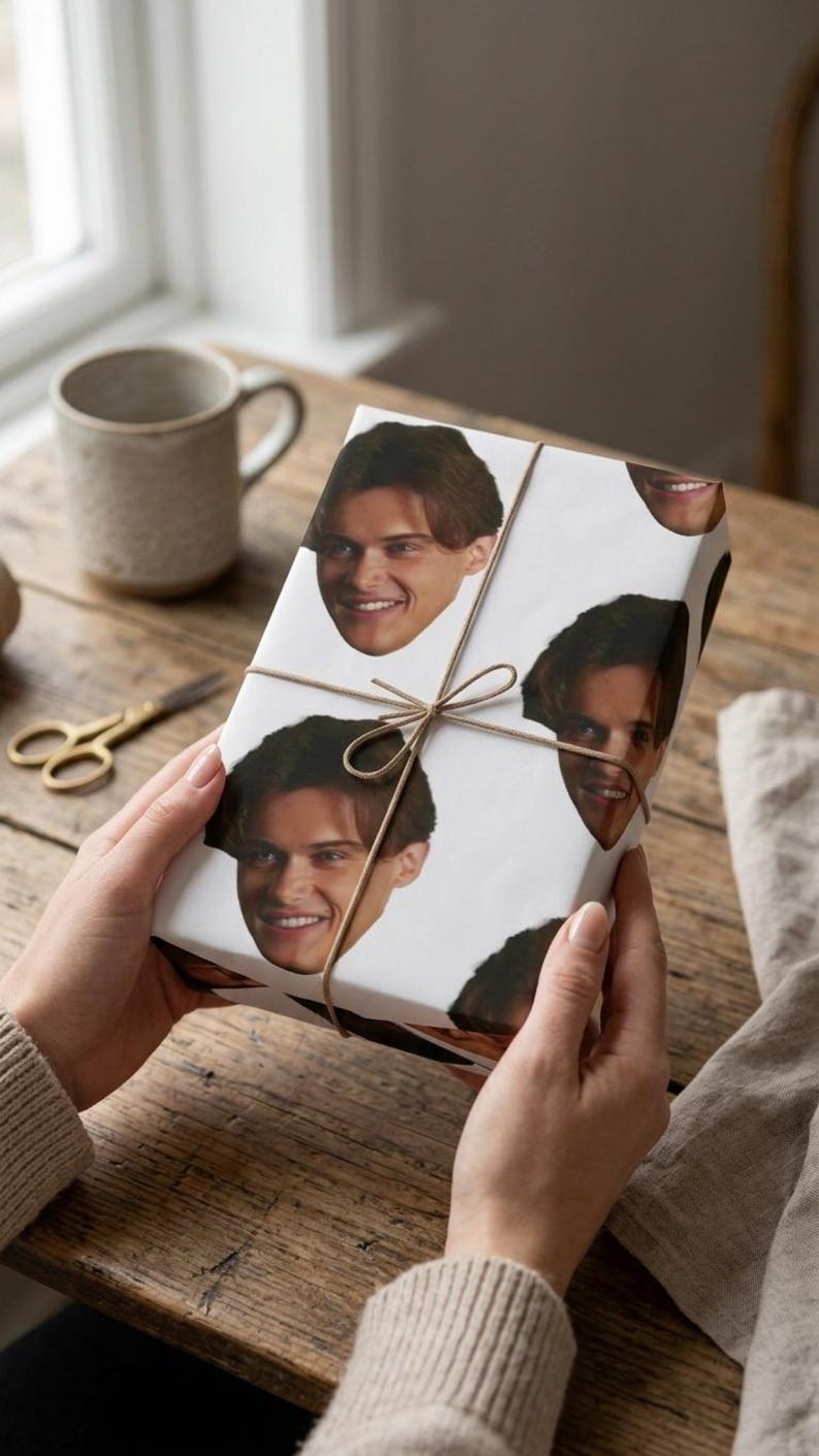 May include: A wrapped gift with a white paper featuring repeating images of a smiling person's face. The gift is tied with twine and held by hands. A pair of gold scissors and a mug are in the background.