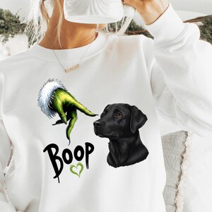 May include: White sweatshirt featuring a Grinch-like green hand, a black Labrador, and the word "Boop" with a green heart. A Santa mug is held up. The sweatshirt is a casual, comfortable style.